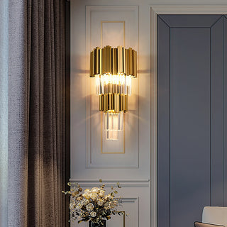 A golden crystal wall sconce is recessed within white wainscoting, flanked by gray curtains and blue-gray paneling, with a white floral bouquet arranged beneath—evoking a luxurious European style.