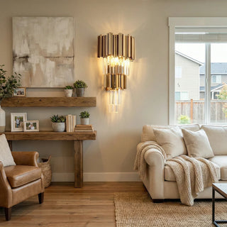 A gold crystal wall lamp is mounted on the wall of a cozy living room, flanked by wooden shelves, greenery, and decorative artwork, and paired with an off-white sofa and a brown leather armchair.