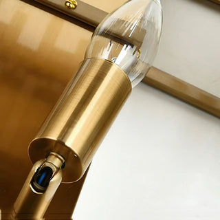 A close-up shot of a wall lamp bulb, showcasing a clear glass E14 candle-style bulb mounted in a gold-tone brass socket, with visible details of the light switch.