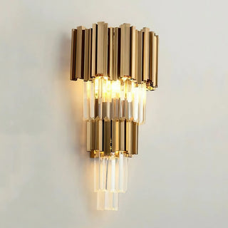 A gold crystal wall lamp, illuminated and mounted against a white wall, features a three-tiered arrangement of metal tubes paired with suspended crystal rods, with warm light radiating outward from the light source.